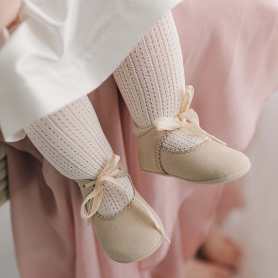 A close-up of a baby’s legs, dressed in Openwork Fancy Tights and wearing beige soft-soled shoes with ribbon ties. The baby is seated, with a pink blanket partially visible in the background, adding a dressy touch to the cozy scene.