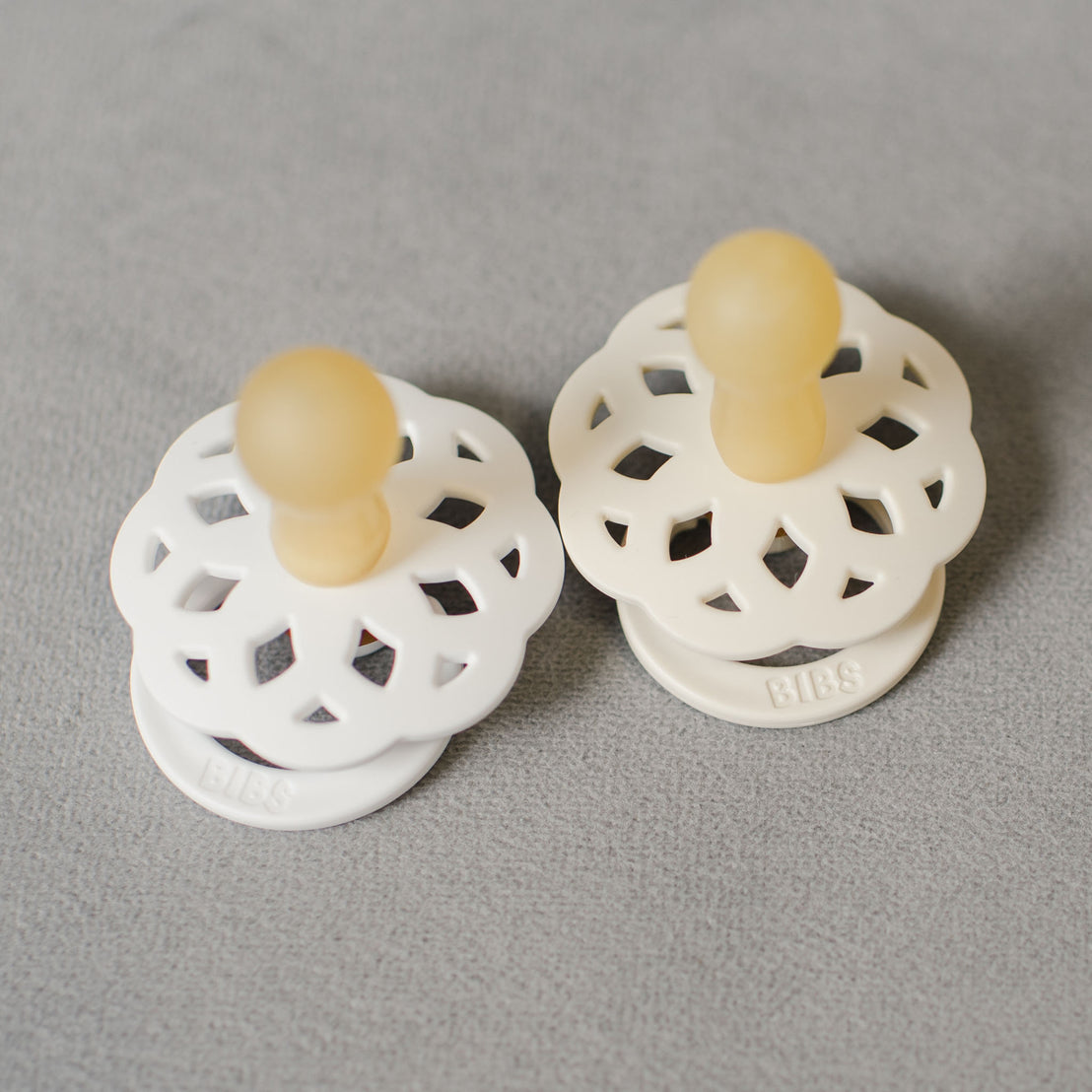 An Aria Pacifier Set in White & Ivory with round, cut-out shields, displayed on a grey textile surface.