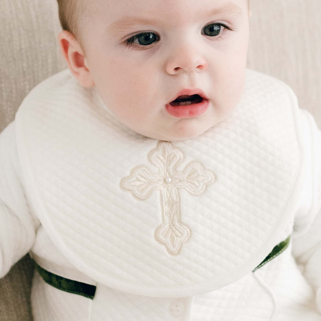 A baby gazes attentively while wearing the Boys Cross Bib, a handmade product crafted in the USA. The bib's white fabric features an embroidered cross design, adding a soft and cozy touch that perfectly complements the calm, indoor setting.