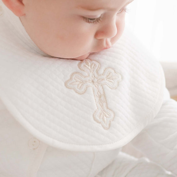 A baby is dressed in a boys cross bib, crafted by hand in the USA, showcasing an elegantly embroidered cross pattern. The child wears a white ensemble, highlighting the bib's detailed embroidery.
