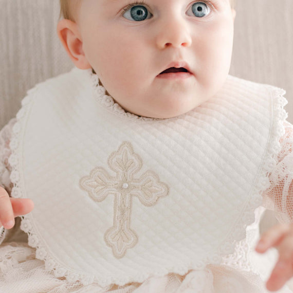 A baby girl is wearing the Girls Cross Bib, a white bib featuring an embroidered decorative cross and a pearl-style button. The background is softly neutral, highlighting her light complexion and blue eyes as she gazes upward, with her outfit's delicate lace details completing the look.