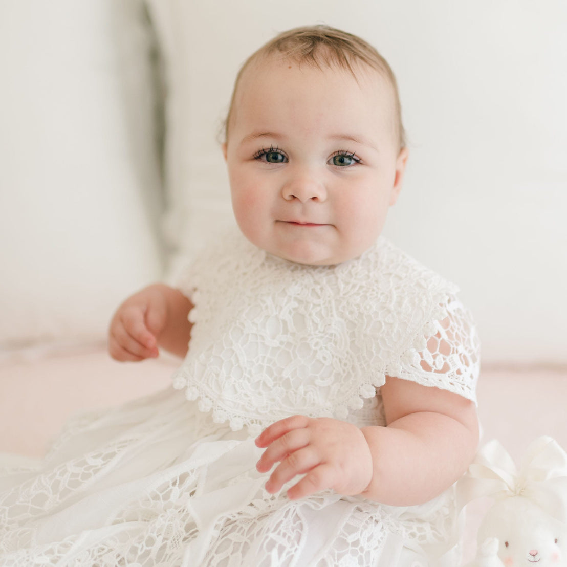 A baby with light skin and rosy cheeks is sitting and wearing a delicate white cotton Grace Lace Bib, reminiscent of a christening gown. The baby has short, light brown hair and blue eyes, gazing towards the camera with a soft smile. There is a plush toy with a bow visible in the lower corner.