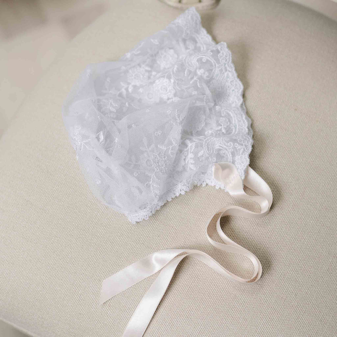 Flat lay photo of the Melissa Sheer Bonnet with lace detailing and pink silk ribbons. Set against the beige surface of a chair. Part of the Baby Beau & Belle Christening Dress Baby Girl Collection. 