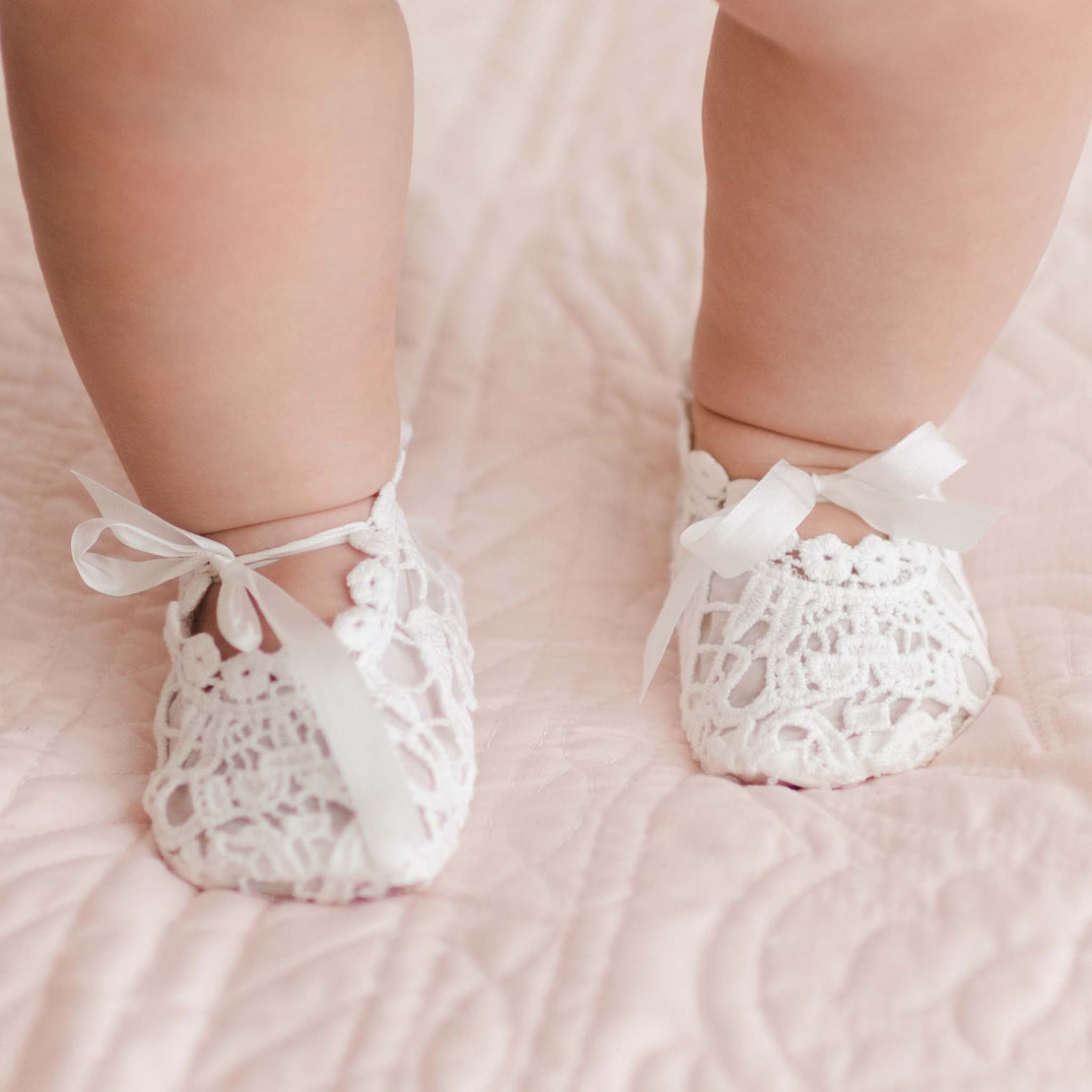 A close-up photo captures a baby's feet adorned in delicate, white Grace Lace Booties with ribbon ties, resting on a soft, pink quilted blanket. The baby's legs are visible while the rest of the body remains out of frame, giving a hint of an exquisite Christening gown crafted from a luxurious silk and cotton blend.