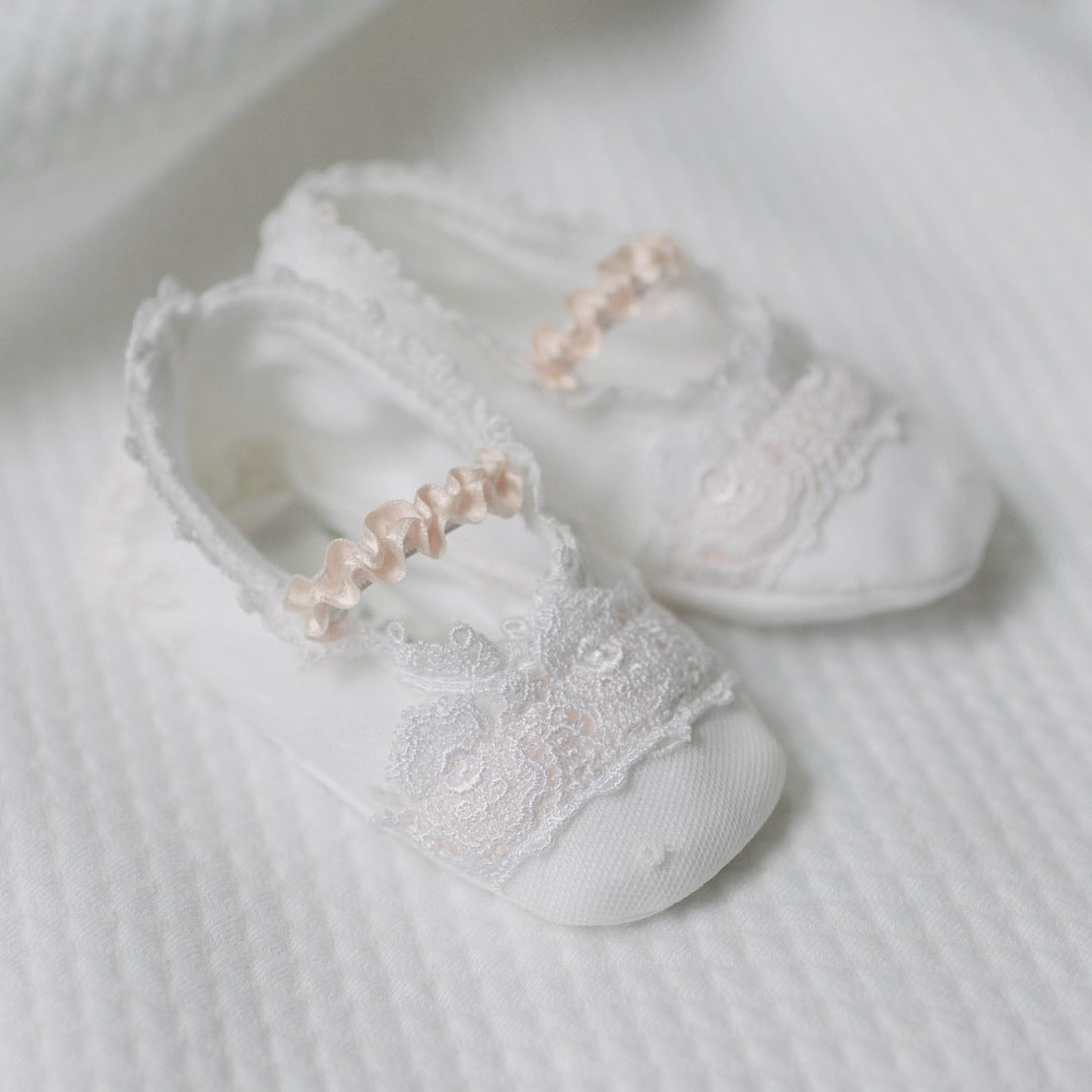 Flat lay photo of the Melissa Booties with lace details and pink ribbons on a white quilt surface. Close up details feature dotted Swiss and floral embroidered pattern netting overlay and white Venice lace covering the top. An accessory to luxury baby clothes. 