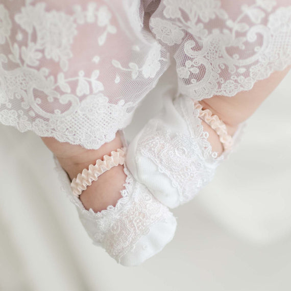 Close-up of a baby's feet dressed in delicate handmade Melissa Booties and white lace-trimmed clothing. The intricate floral patterns of the Venice lace create an elegant and soft appearance, with the 100% pima cotton ensuring comfort. The blurred background draws focus to the baby's feet.