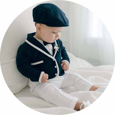 A baby sits on a white bed dressed in a navy blue and white outfit, complete with a matching cap and bow tie, looking downward. The room is softly lit, creating a serene atmosphere.