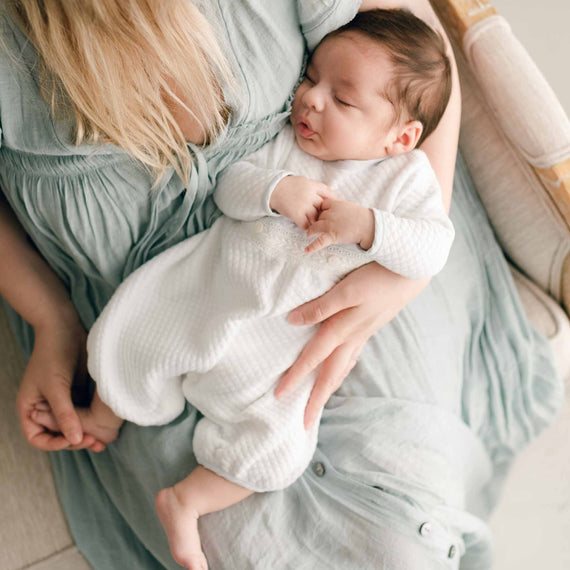 A sleeping baby cradled in a mother's arms, wearing the Harrison Newborn Romper made of textured white fabric with lace detailing at the waist.
