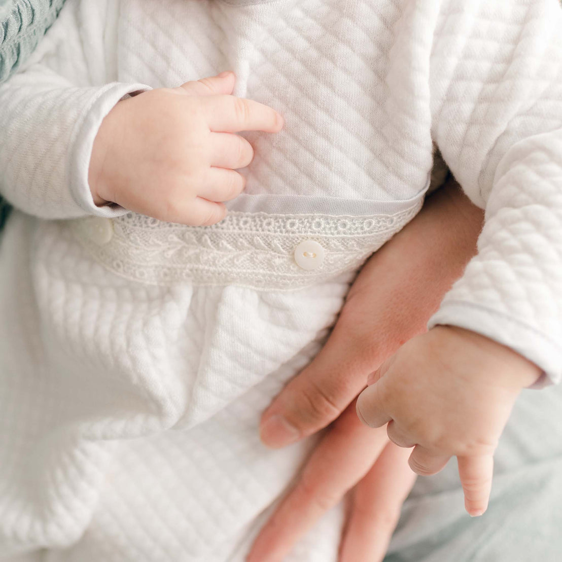 A close-up of a baby’s hands resting on the textured fabric and lace waistline of the Harrison Newborn Romper.