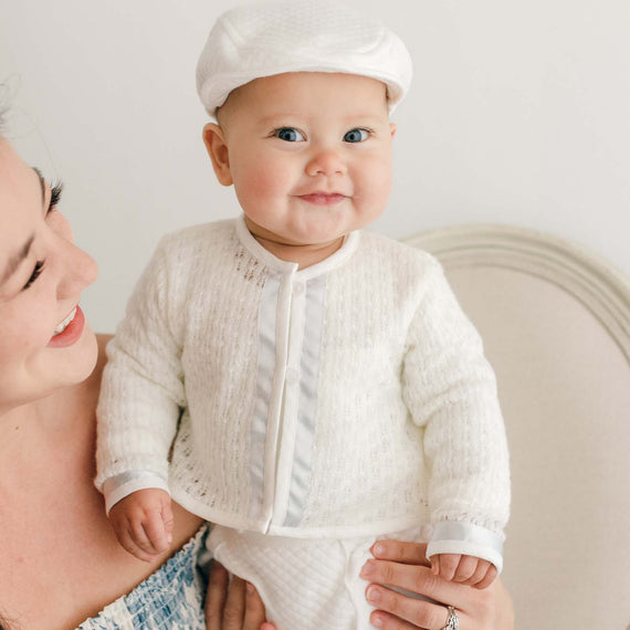 Smiling baby boy standing on a chair with his mother nearby. He is wearing the Owen 3-Piece Suit, including the sweater, pants, and onesie