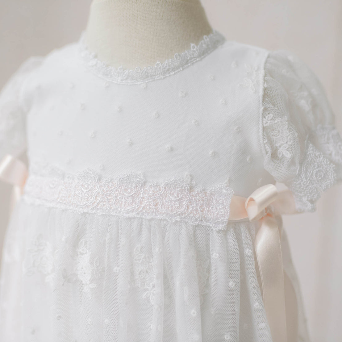 Close-up of the Melissa Christening Gown bodice on a mannequin against a neutral background. Details show the the white Venice lace trim and pale pink silk ribbons that adorns the bodice. Part of the Baby Beau & Belle Christening Gowns Collection.