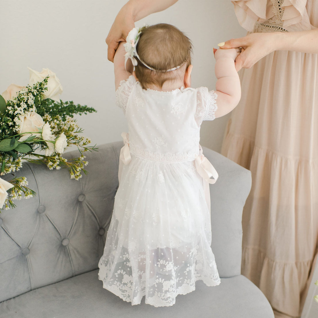 A baby in a Melissa Romper Dress is held up by her mother, who is adjusting a floral headband on her head, in a softly decorated room with flowers nearby.