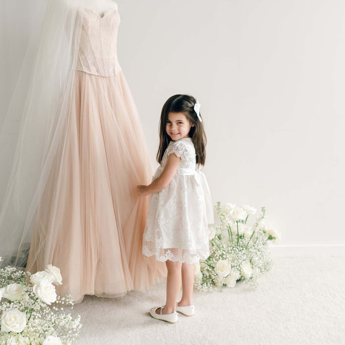 Girl wearing the Penelope Lace Dress, holding blush fabric with white floral arrangements in the background.