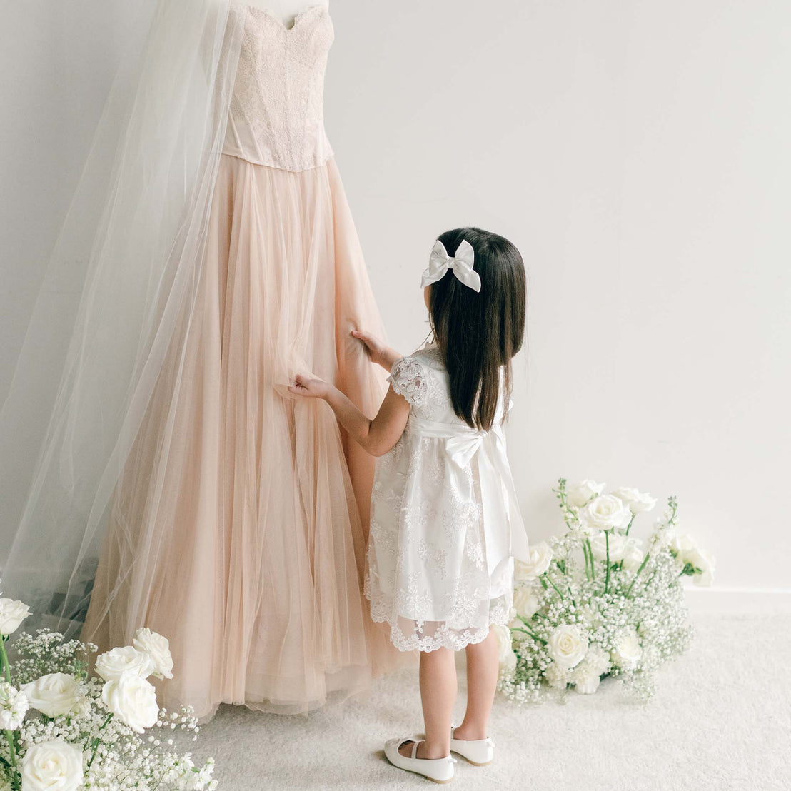 Back view of the Penelope Lace Dress, showing the satin bow tied at the waist as the girl looks at a blush gown.