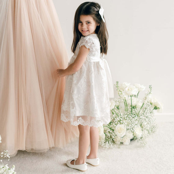 Little girl wearing the Penelope Lace Dress, standing beside a blush gown and white floral arrangement.