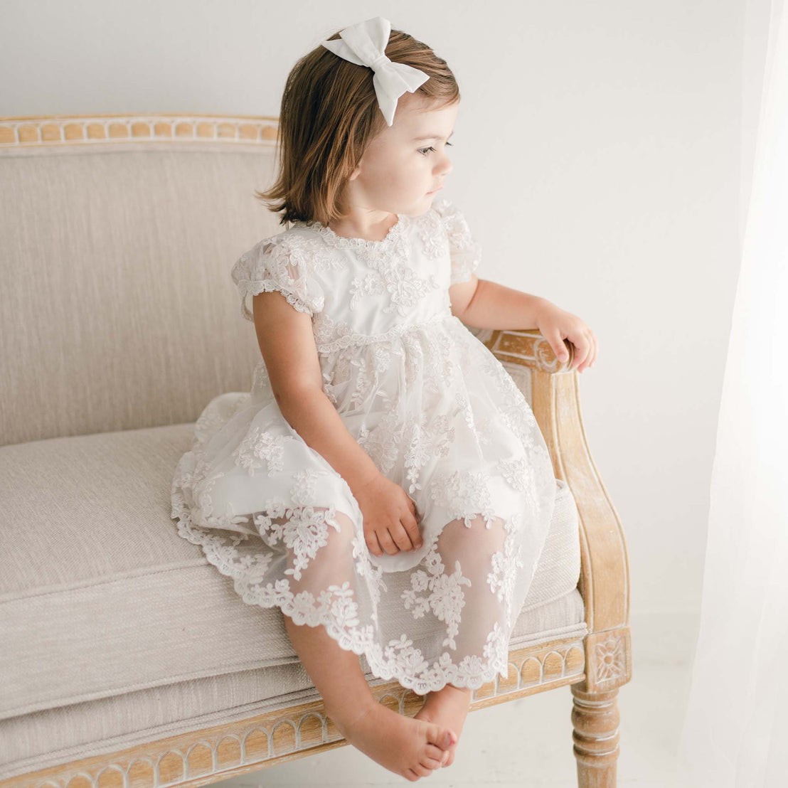 A young girl in a delicate Penelope Lace Dress adorned with embroidered lace sits on an elegant beige couch. She gazes toward a bright window, with soft light illuminating the room. Her feet are bare, and her expression is calm and serene.