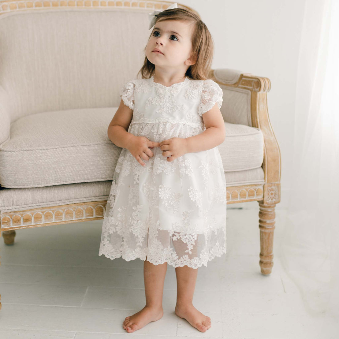 A young child stands barefoot on a light-colored floor, wearing the elegant Penelope Lace Dress, which is beautifully adorned with embroidered lace. The child looks upward with a neutral expression, while behind them sits an ornate, light-colored armchair.