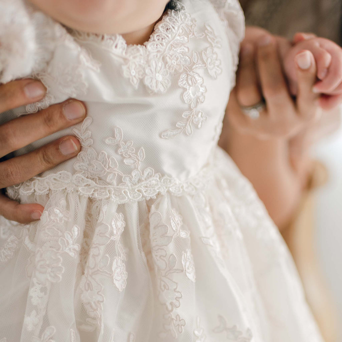 A close-up of a child in the Penelope Lace Dress, adorned with intricate floral embroidery. An adult, partially visible, holds the child's hand gently, creating a warm and tender scene.