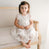 A young child with brown hair sits on an ornate cream-colored sofa, wearing the Penelope Lace Dress, a white christening gown adorned with embroidered lace. A delicate white bow graces her hair, while the background remains a soothing, neutral tone.