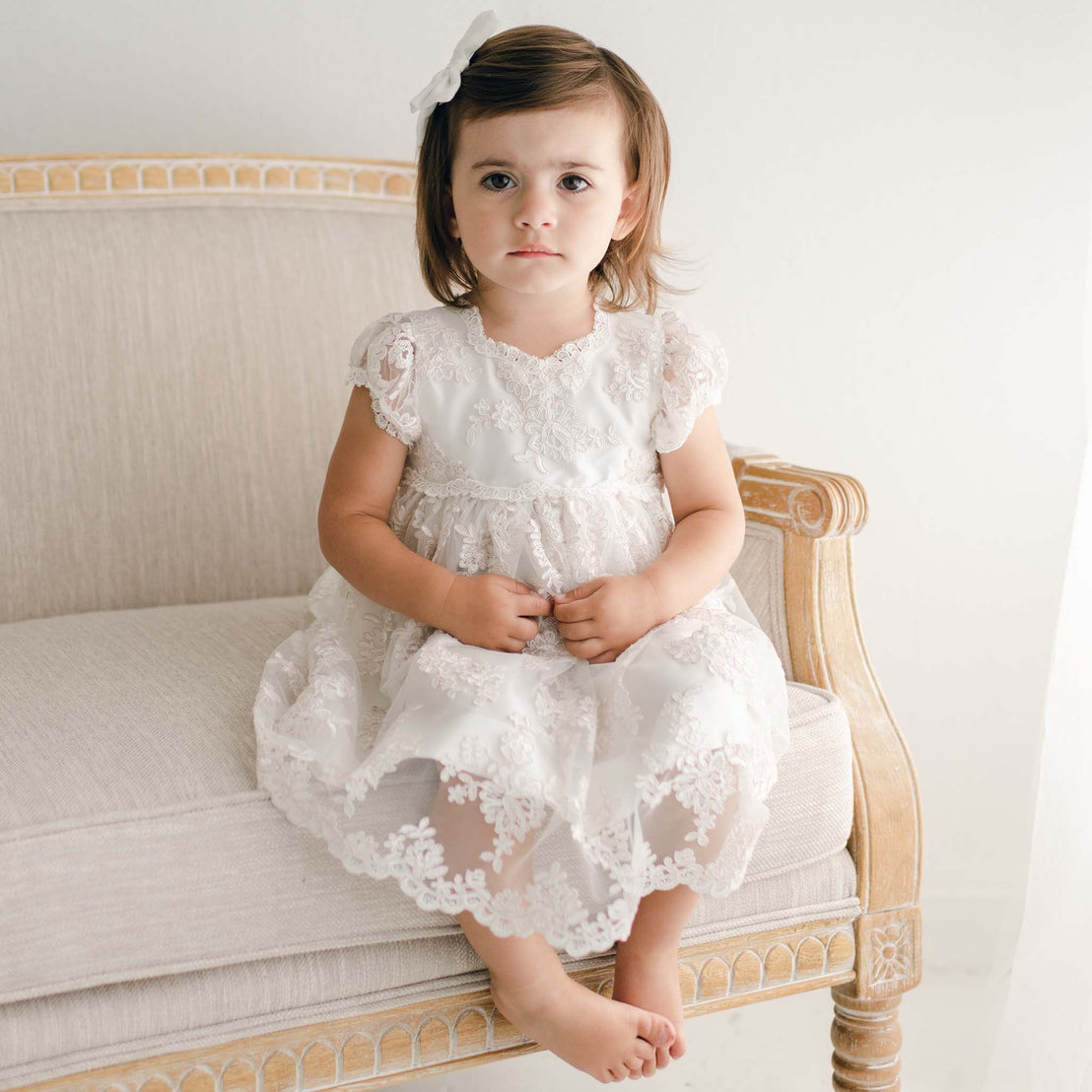 A young child with brown hair sits on an ornate cream-colored sofa, wearing the Penelope Lace Dress, a white christening gown adorned with embroidered lace. A delicate white bow graces her hair, while the background remains a soothing, neutral tone.