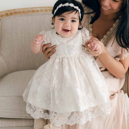 A woman smiles at a baby adorned in a Penelope Lace Dress with a matching headband. The baby, seated on a beige sofa and clasping the woman's hands, radiates joy. Both are dressed in light-colored clothing, capturing a heartwarming moment of pure happiness.