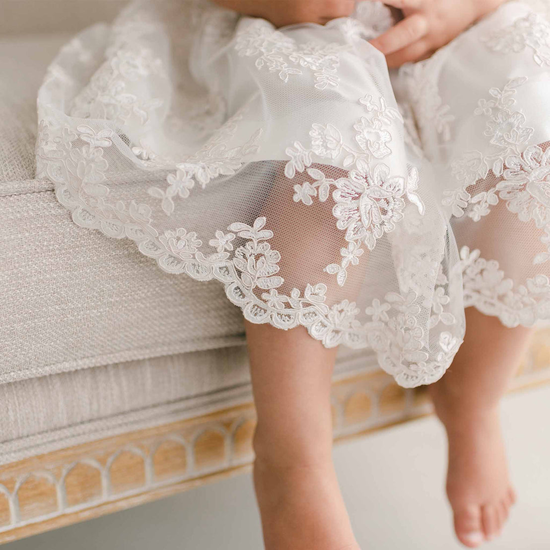 Close-up of a child's feet and legs in the Penelope Lace Dress, showcasing intricate floral patterns along the hem. The white embroidered lace of the Penelope Dress is elegantly draped as the child sits on a textured light-colored couch.