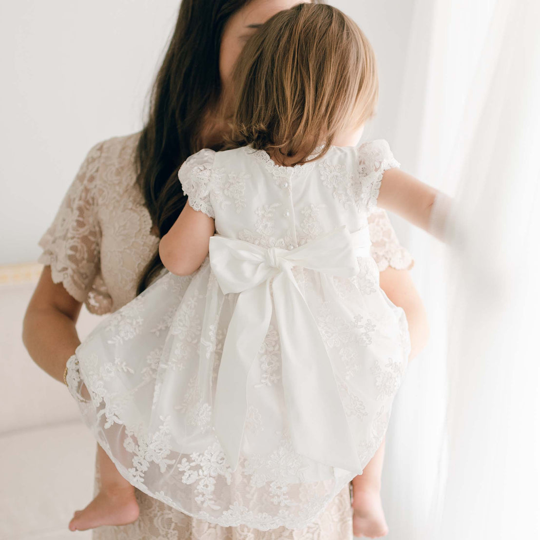 A woman cradles a small child dressed in the Penelope Lace Dress, a silk christening gown embellished with intricately embroidered lace and a large bow. Partially visible, the woman adds to the softly lit scene, creating a serene and tender moment.