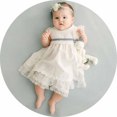 A baby wearing a white lace dress and a floral headband lies on a soft, light blue surface. The baby holds a small stuffed toy rabbit, looking upward with a calm expression.