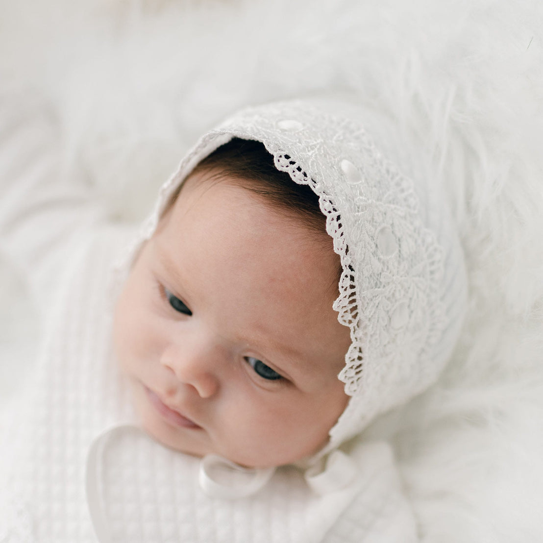 A newborn baby wearing a Madeline Quilted Newborn Bonnet, an heirloom piece, lies on a soft, fluffy white blanket, looking thoughtfully to the side.