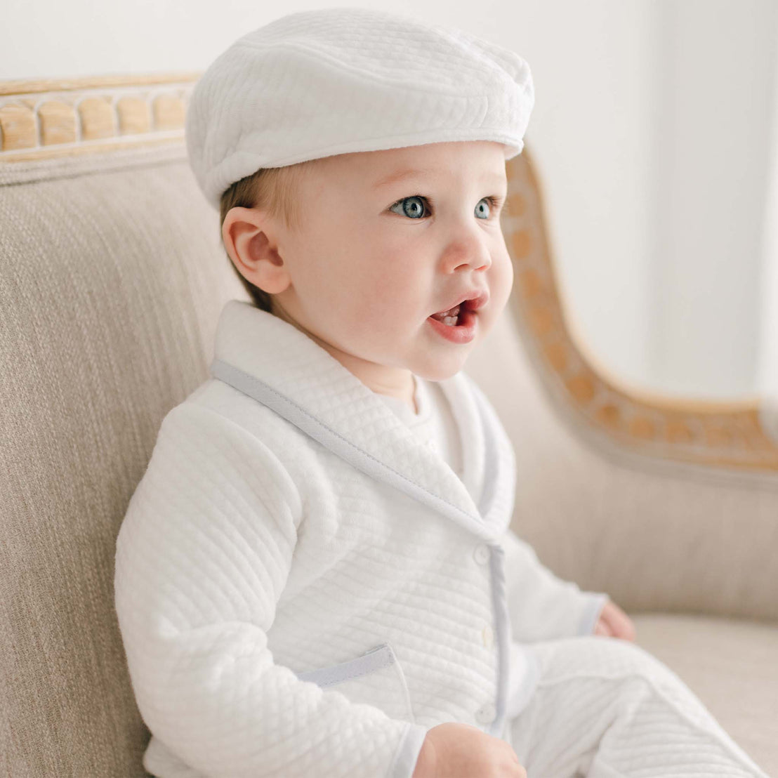 Side view of a baby wearing the Harrison Quilted Newsboy Cap, paired with the matching quilted pants suit, sitting on a beige cushioned chair.