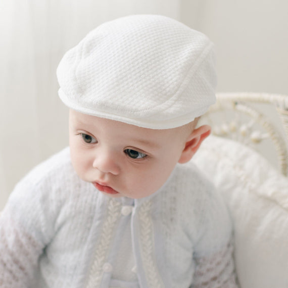 A baby wearing the Harrison Textured Newsboy Cap, made of soft quilted fabric in white, sitting on a vintage-inspired chair.