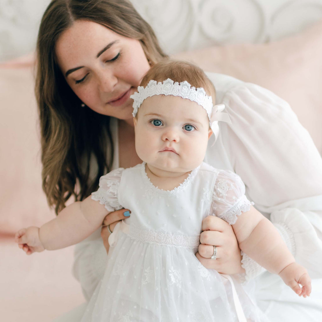 A woman with long brown hair, wearing a white blouse, is gently holding a baby dressed in a white Venice lace dress and the Melissa Headband, which is adorned with a pink silk ribbon. The baby has blue eyes and looks directly at the camera, while the woman smiles softly, looking at the baby.