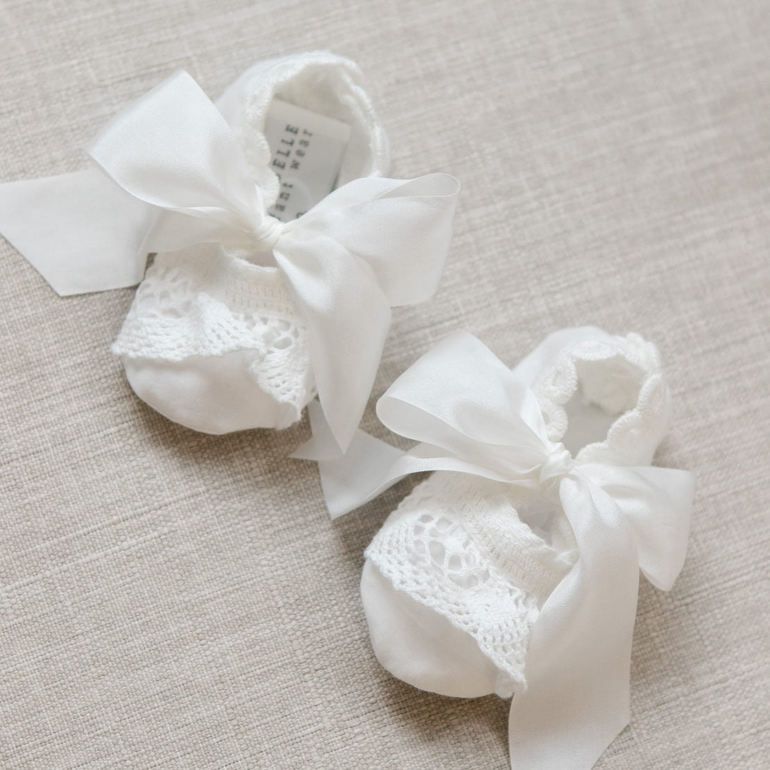 Pair of white baby booties with lace details and ribbons on a beige surface