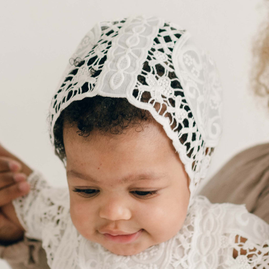 Close-up of a baby girl wearing the Adeline Lace Bonnet, featuring intricate ivory lace with delicate floral detailing. The bonnet sits softly on the baby’s head, framing the face with its unique openwork design.