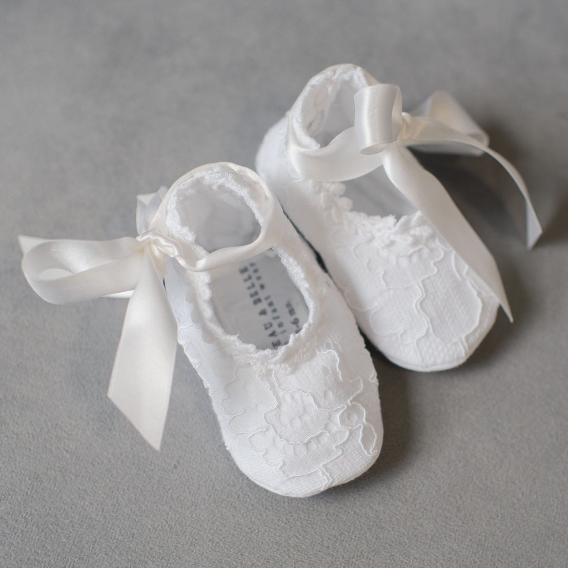 Aria Lace Booties, featuring elegant lace detailing and satin bows, are the ideal accessory to complete a baptism outfit, beautifully displayed on a soft gray background.