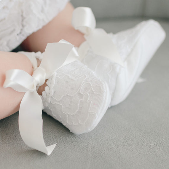 A close-up of a baby girl wearing the Aria Lace Booties, crafted in delicate white lace and adorned with large satin bows, as she rests on a soft gray surface. She is dressed in matching lace attire, creating an enchanting baptism outfit.