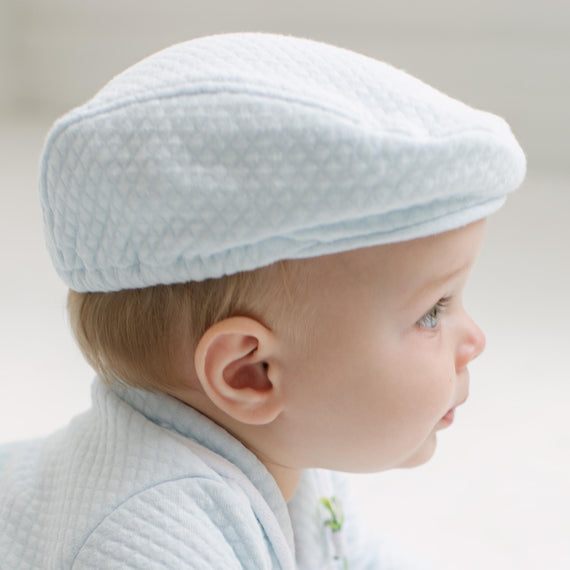 A close-up side profile of a baby wearing a light blue textured flat cap and matching light blue quilted clothing against a soft, light gray background.