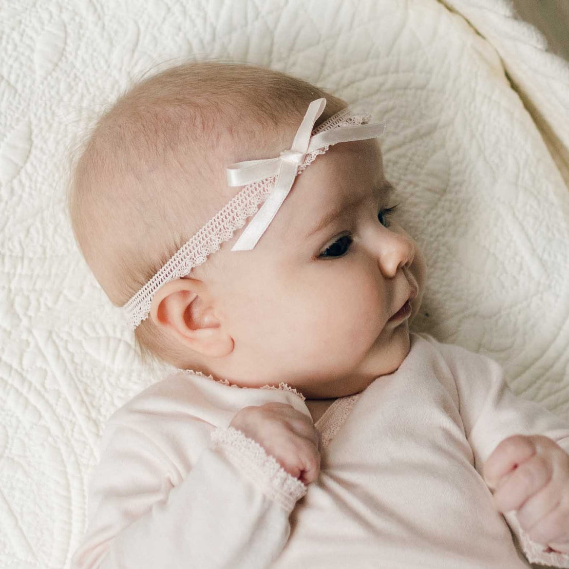 Close up of a newborn baby on a white quilt wearing an Ava Lace Headband with matching Ava Layette Coming Home Outfit. Details show blush silk ribbon bow with matching stretch lace.