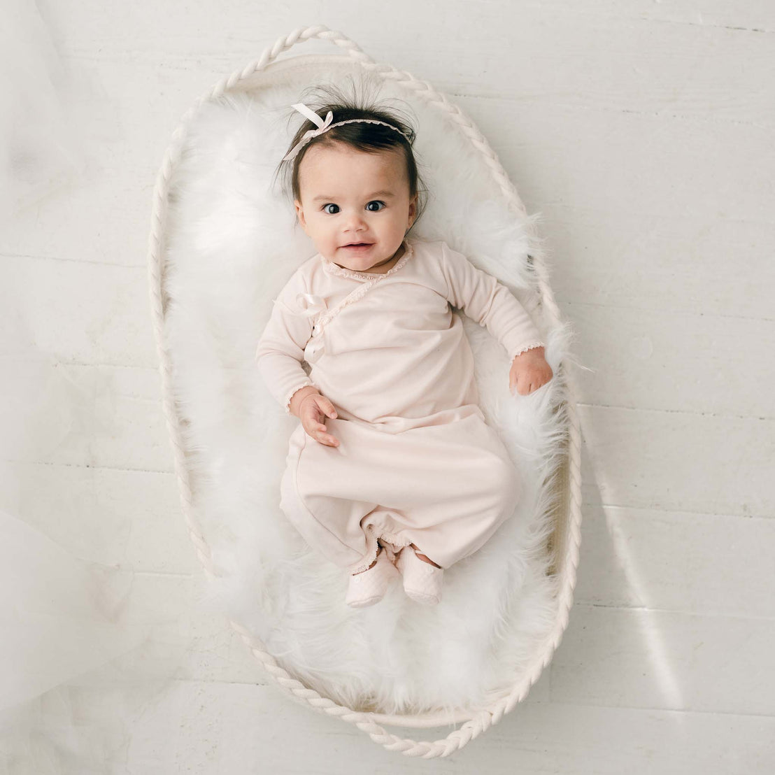 A newborn girl with dark hair wears the Ava Headband and matching Ava Newborn Layette and Ava Quilted Booties lies in an oval-shaped basket lined with soft white fur and looks upwards with a slight smile