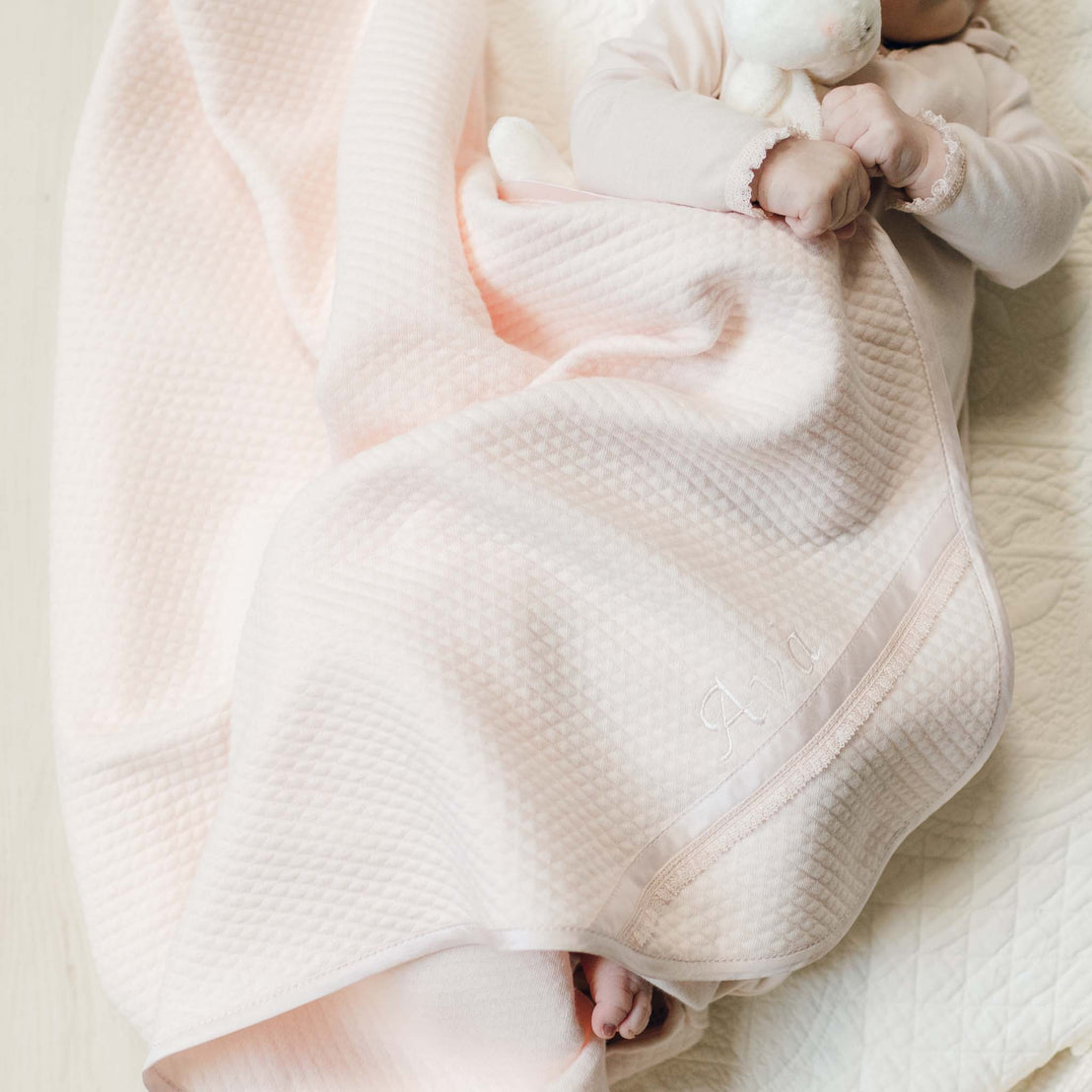 Newborn baby wrapped in the blush pink Ava Personalized Quilt Blanket. Corner detail of the blanket features the custom embroidered name "Ava" along the silk dupioni and lace trim. Part of the Baby Beau & Belle Baptism Gift From Godparents Collection.