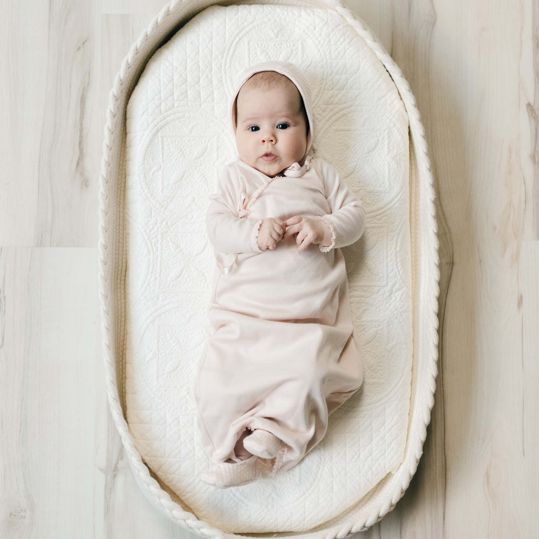 Newborn baby in the plush pink Ava Newborn Layette Coming Home Outfit with matching quilt bonnet lying in a beige quilt crib set against a wood floor. A perfect coming home outfit for baby.