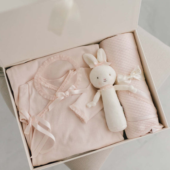 A gift box containing the Ava Newborn Gift Set, including the Ava Newborn Layette, Ava Headband, a plush Bunny Chime, and an Ava Personalized Blanket. Set against a beige chair and white backdrop. A unique coming home outfit for baby gift for godparents.