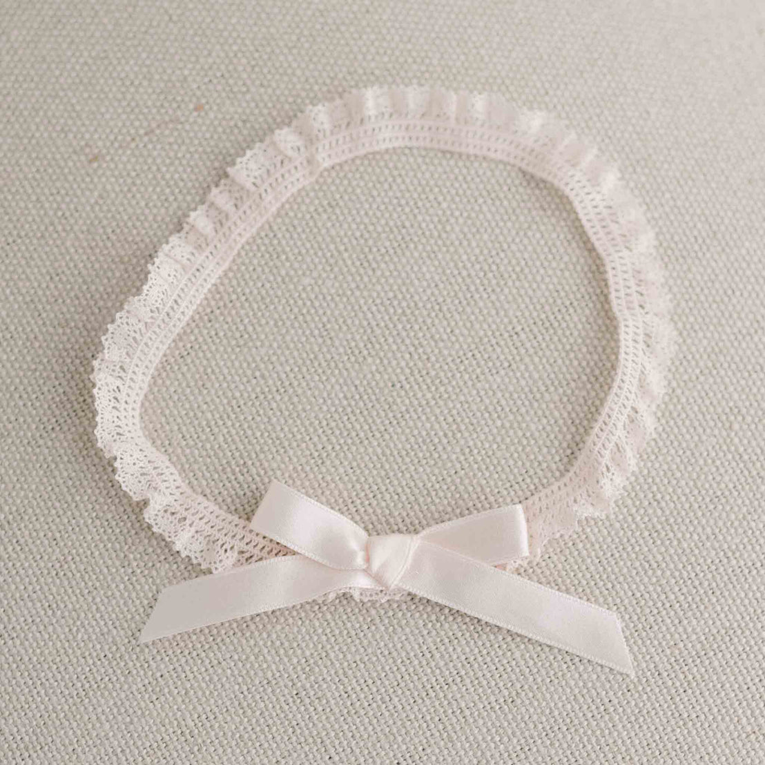 A delicate Ava Headband made from a blush pink lace with a matching silk ribbon bow, placed on a light grey textured background. 