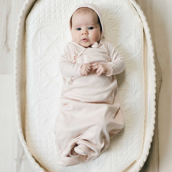 Newborn baby dressed in the blush pink cotton Ava Layette baby girl coming home outfit with quilt bonnet, and lying in a beige crib.