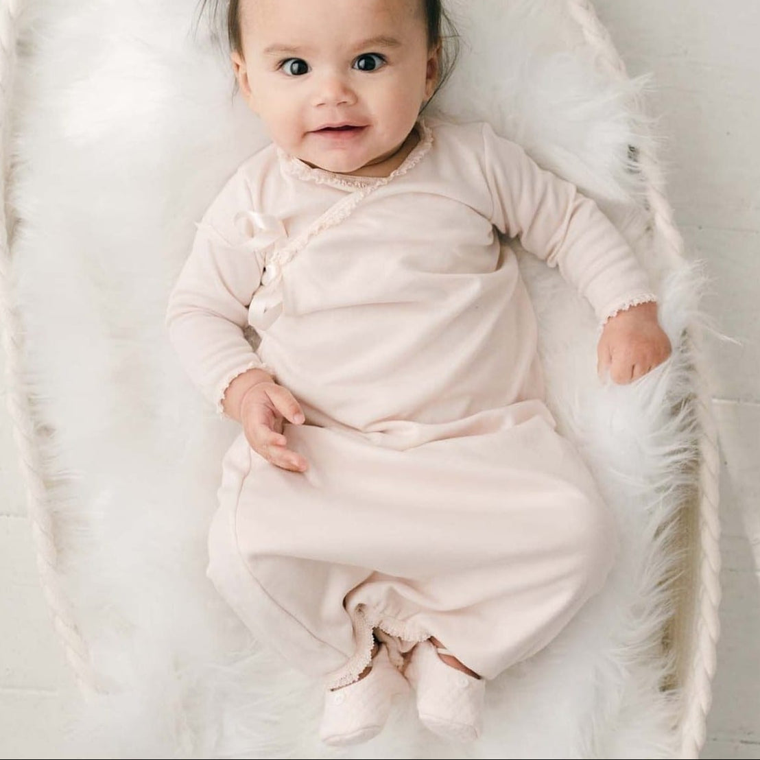 A baby wearing the wearing the Ava Newborn Layette & Headband, a pima cotton layette in a shade of blush pink and matching blush ribbon bow headband, lies smiling in a white fluffy blanket.