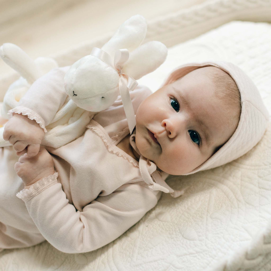 Close-up photo of a newborn holding a white plush bunny toy with a pink bow. The baby girl is dressed in the blush pink Ava Layette with matching quilt bonnet. Part of the Baby Beau & Belle Bring Baby Home Outfit Collection.