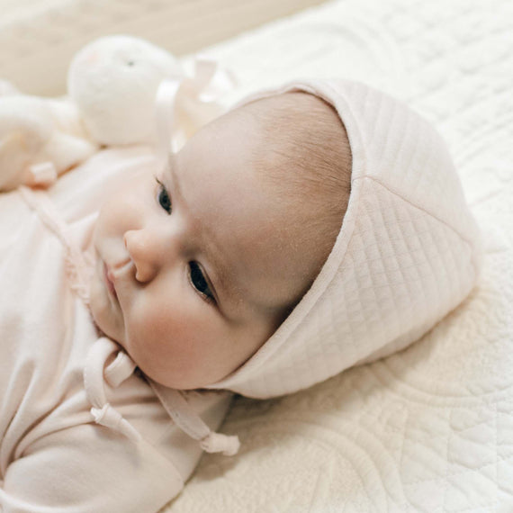 Close-up photo of a newborn in the blush pink Ava Quilt Bonnet laying on a white quilt with a plush bunny beside her. Part of the Baby Beau & Belle Baby Girl Coming Home Outfit Collection.