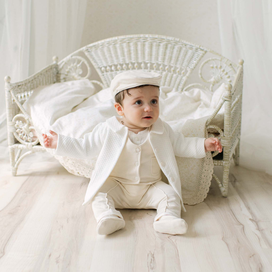Baby boy in the Braden Coat Suit and matching Newsboy Cap sitting on a wooden floor with a white wicker chair in the background. Part of Baby Beau & Belle's luxury baby clothes.
