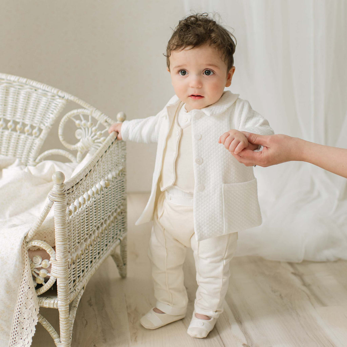 Baby boy in a Braden Coat Suit and matching Vest Suit standing next to a wicker chair in a bright room. Part of the Baby Beau & Belle Baby Boy Suit Collection.