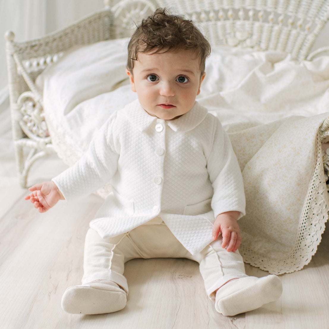 Baby in the Braden Coat Suit sitting on a wooden floor with a white wicker chair and patterned blanket in the background. Perfect choice for high end baby boy suits. 